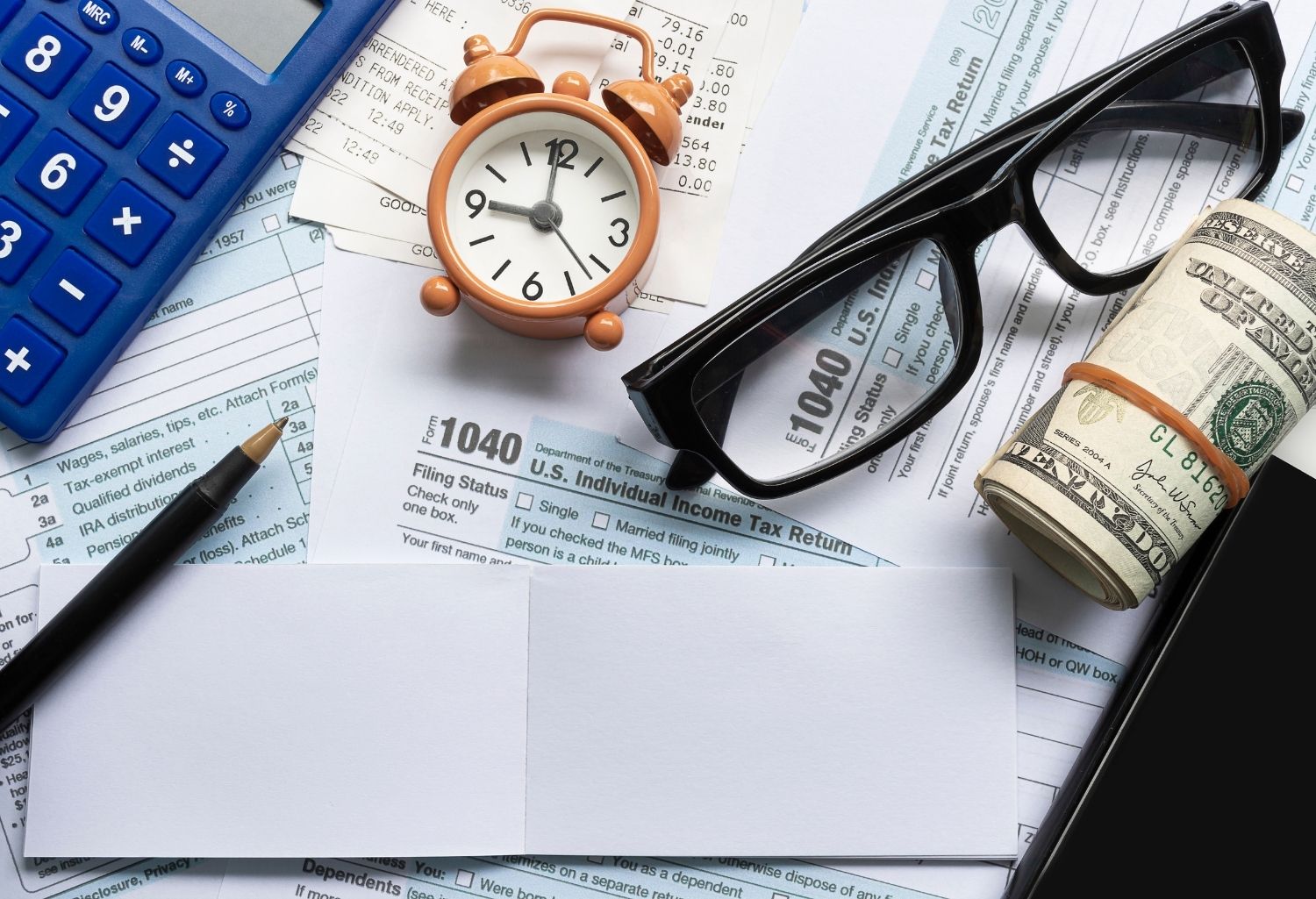 A pen a notepad, rolled up hundred dollar bills, glasses, a clock, a calculator, receipts, and bunch of 1040 forms representing Bank Levy.